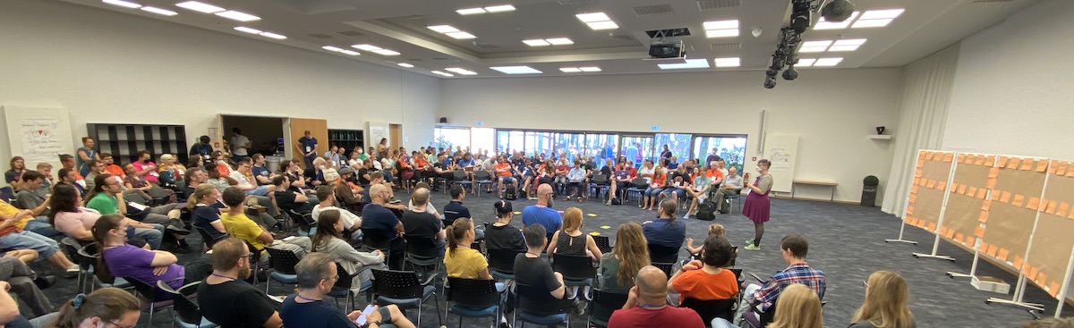 Opening session in a large room with ~200 people. The facilitator stands in front of the group. Behind the facilitator, the board for the Open Space marketplace is visible